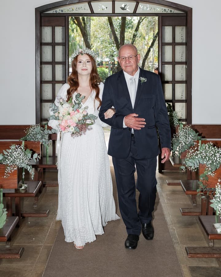 Noiva entrando na capela de braço dado com o pai, entre bancos decorados com flores